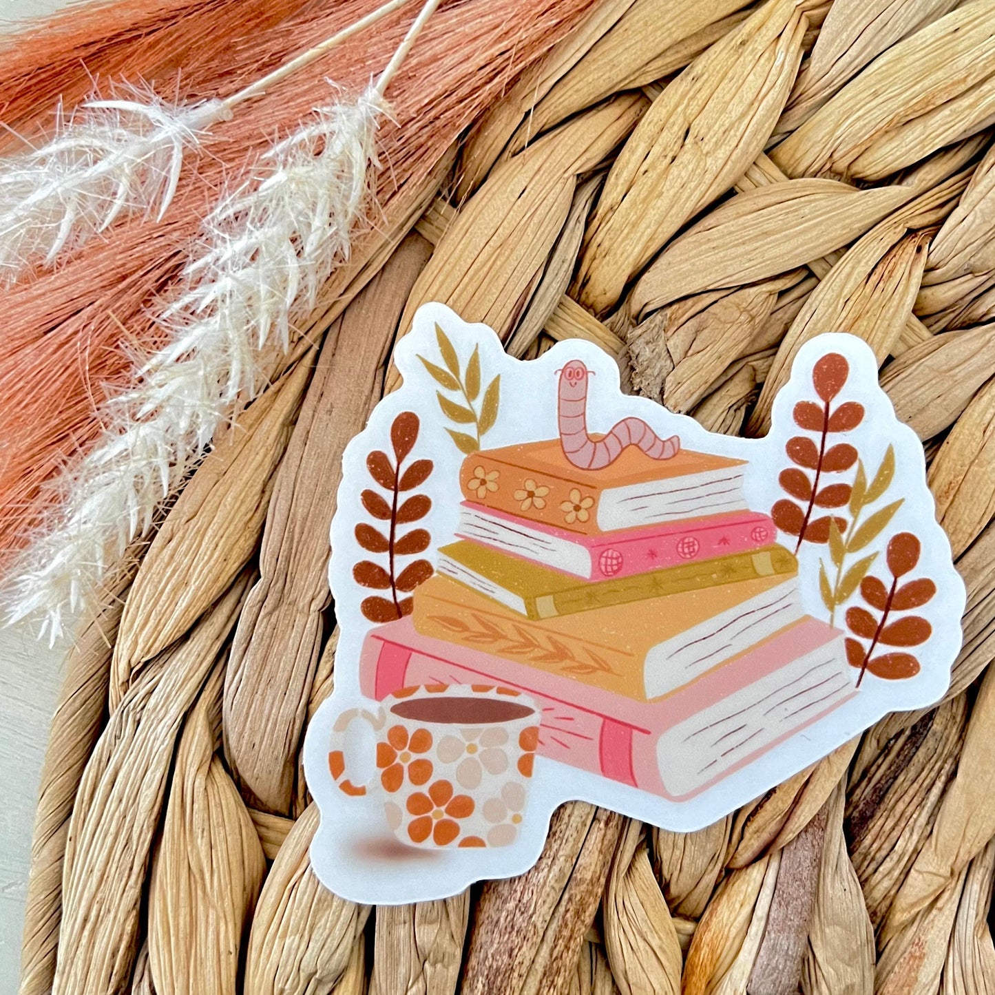 Sticker of books and a mug and worm sitting on top of the books on a woven surface