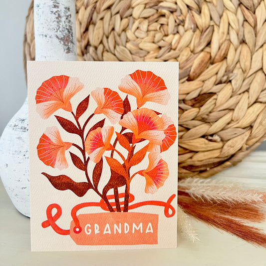 Card with floral design and 'Grandma' text, placed on a woven mat with a white vase and dried plant.