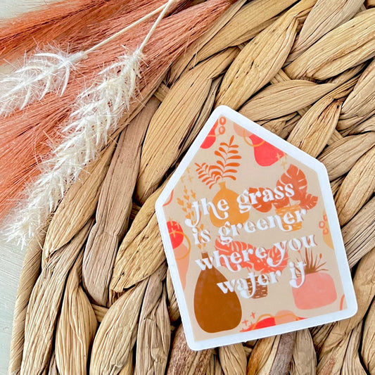 Decorative sticker with text saying "the grass is greener where you water it" on a woven surface with dried leaves.