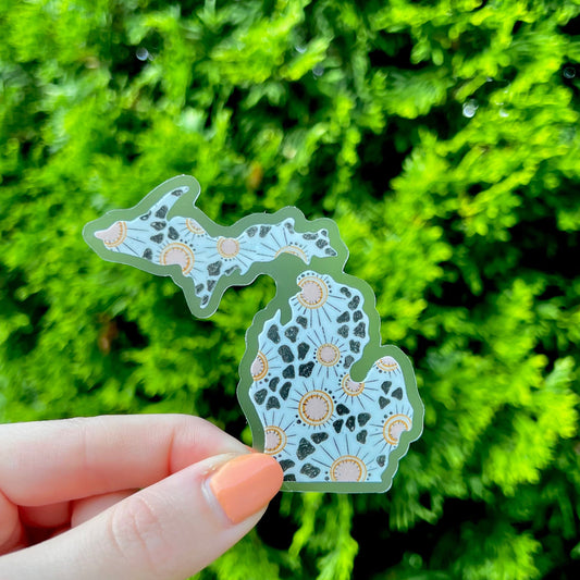 Hand holding a decorative Michigan-shaped sticker in front of green foliage