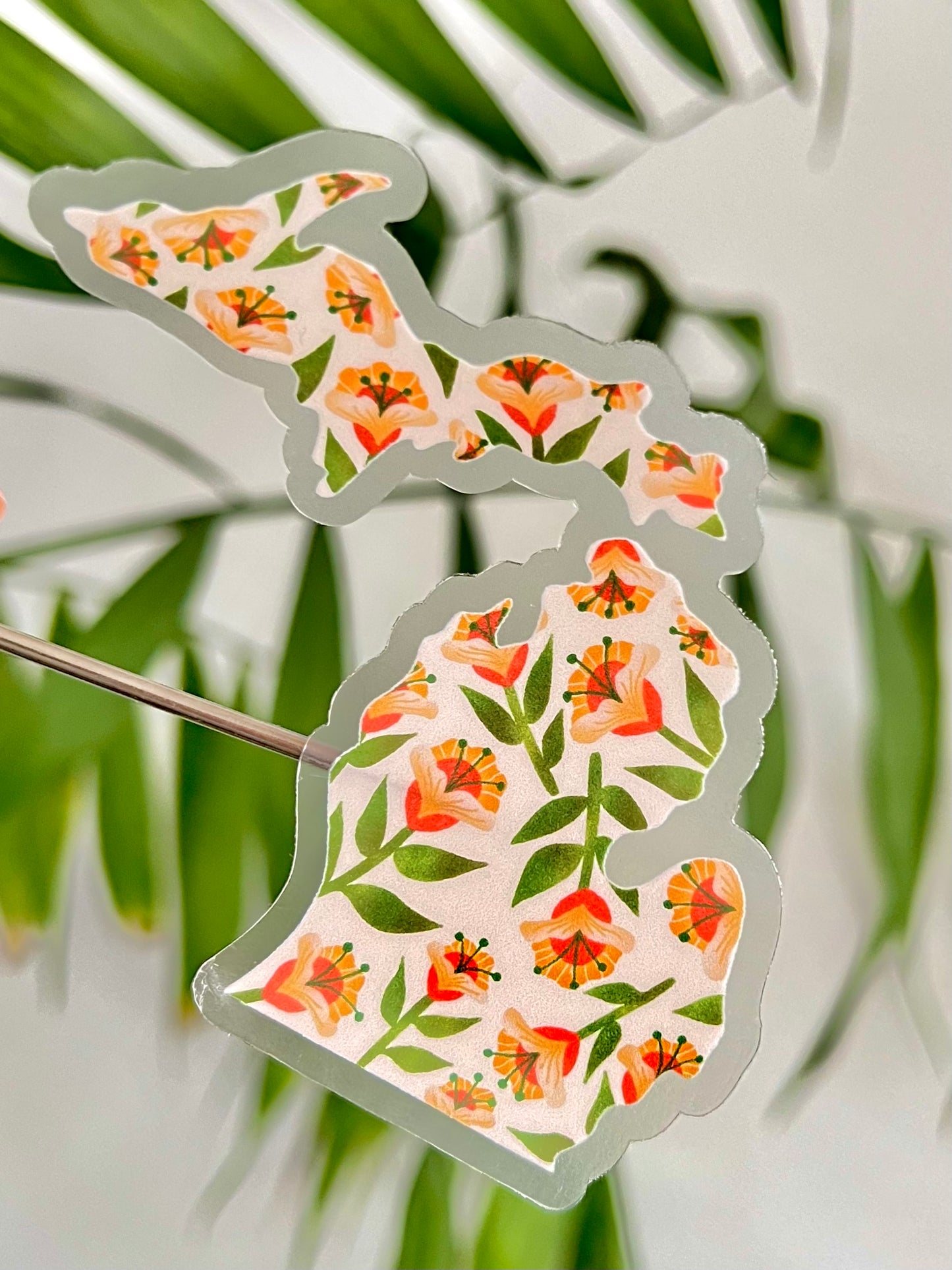 Sticker shaped like the state of Michigan with a illustrated floral poppy design hel up against a blurred natural background
