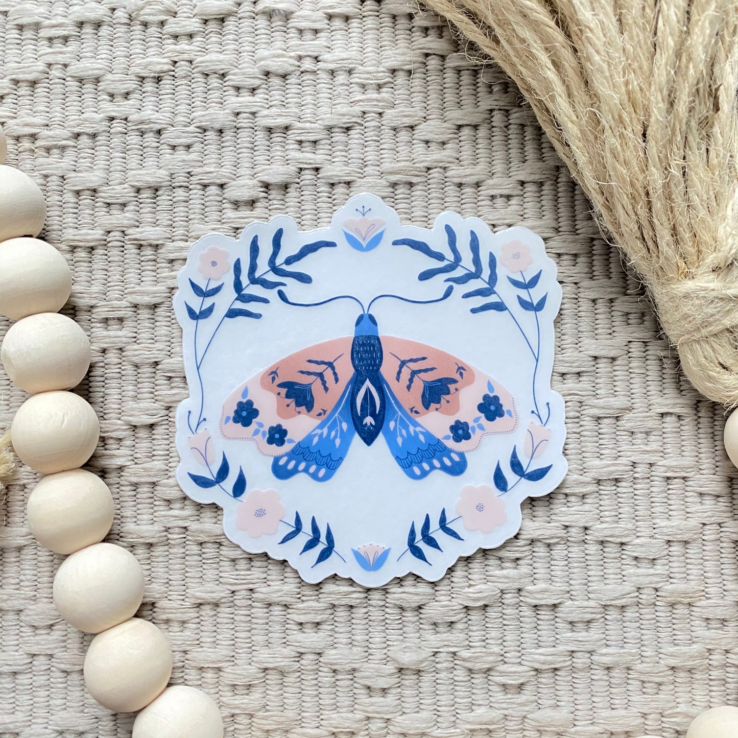 Decorative sticker with a blue and pink moth design on a textured surface with wooden beads and twine.