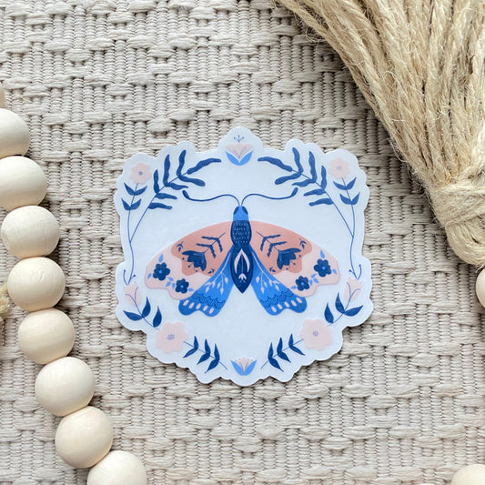 Decorative sticker with a blue and pink moth design on a textured surface with wooden beads and twine.