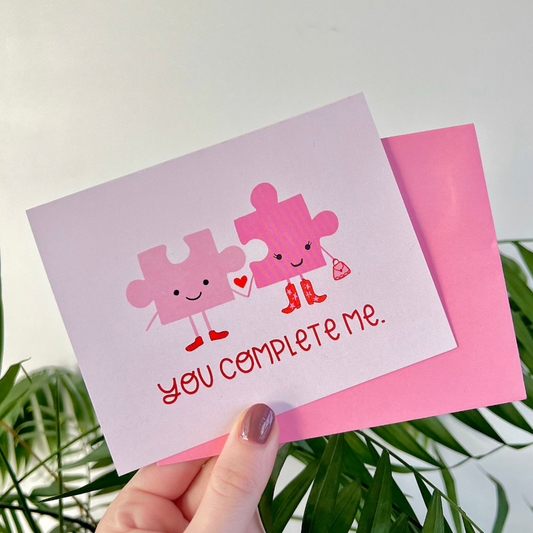 Greeting card with two pink puzzle pieces and text 'You Complete Me' held by a hand with a pink envelope.