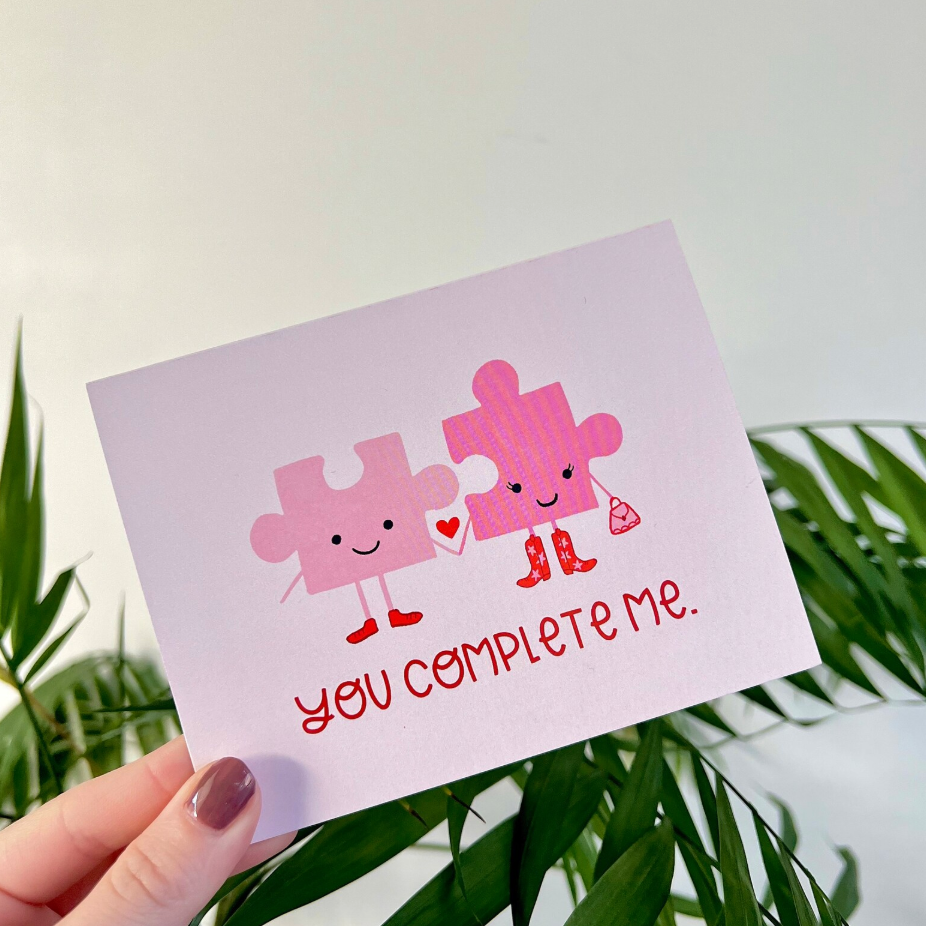 Card with two smiling puzzle pieces holding hands and text 'You complete me' against a light purple background.
