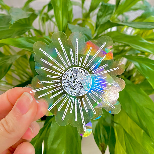 Hand holding a colorful, sun-shaped holographic suncatcher sticker in front of green leaves