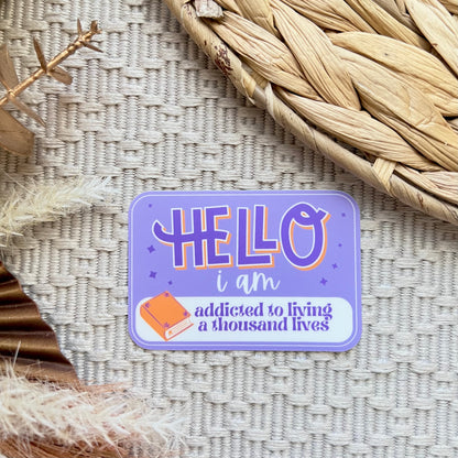 Purple sticker with text 'HELLO i am addicted to living a thousand lives' on a textured surface