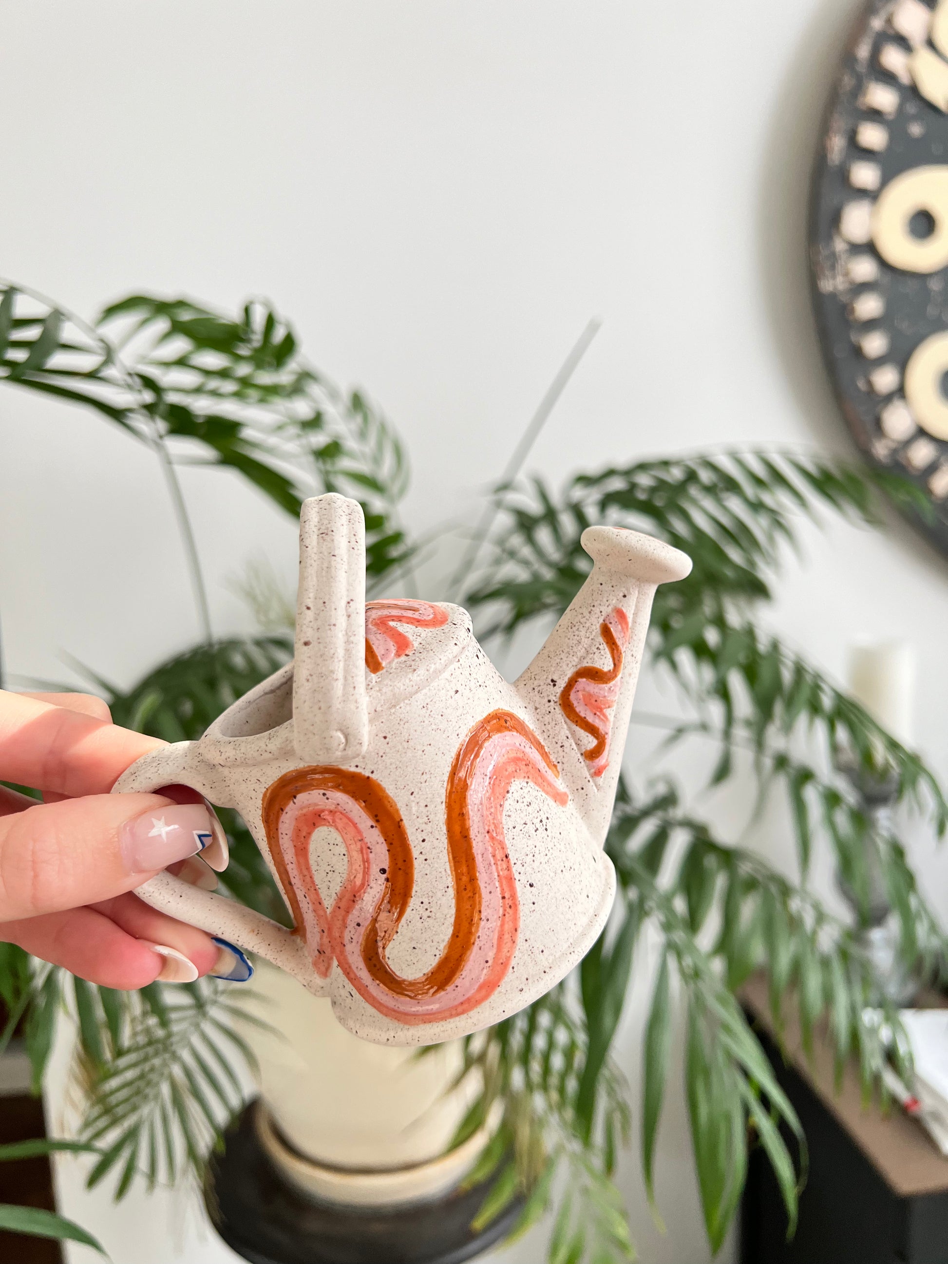 Hand holding a ceramic watering can planter with decorative patterns against a blurred indoor background