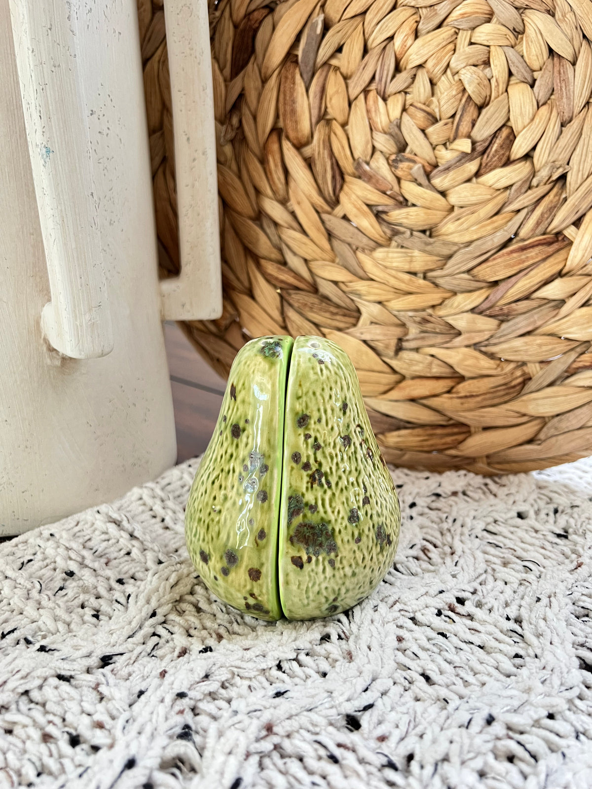 Green ceramic avocado-shaped object on a textured surface with woven basket in the background