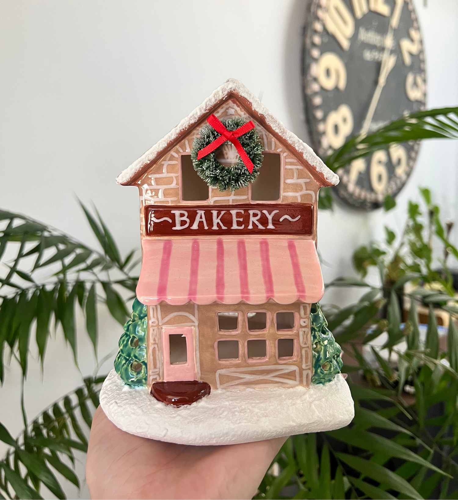 Decorative bakery ceramic house held in a hand with plants and a clock in the background.