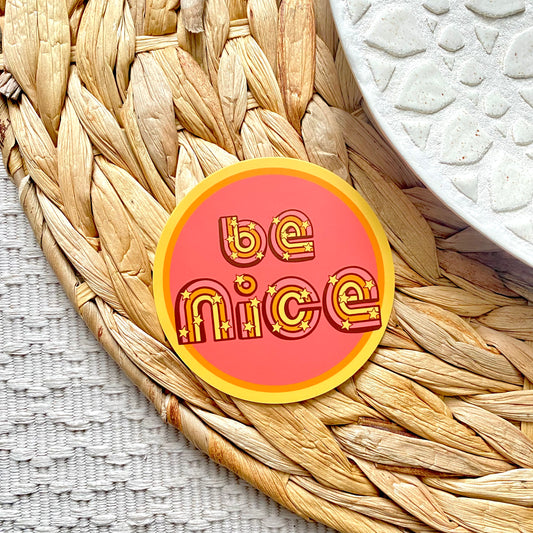 Round pink sticker with 'be nice' text on a woven mat