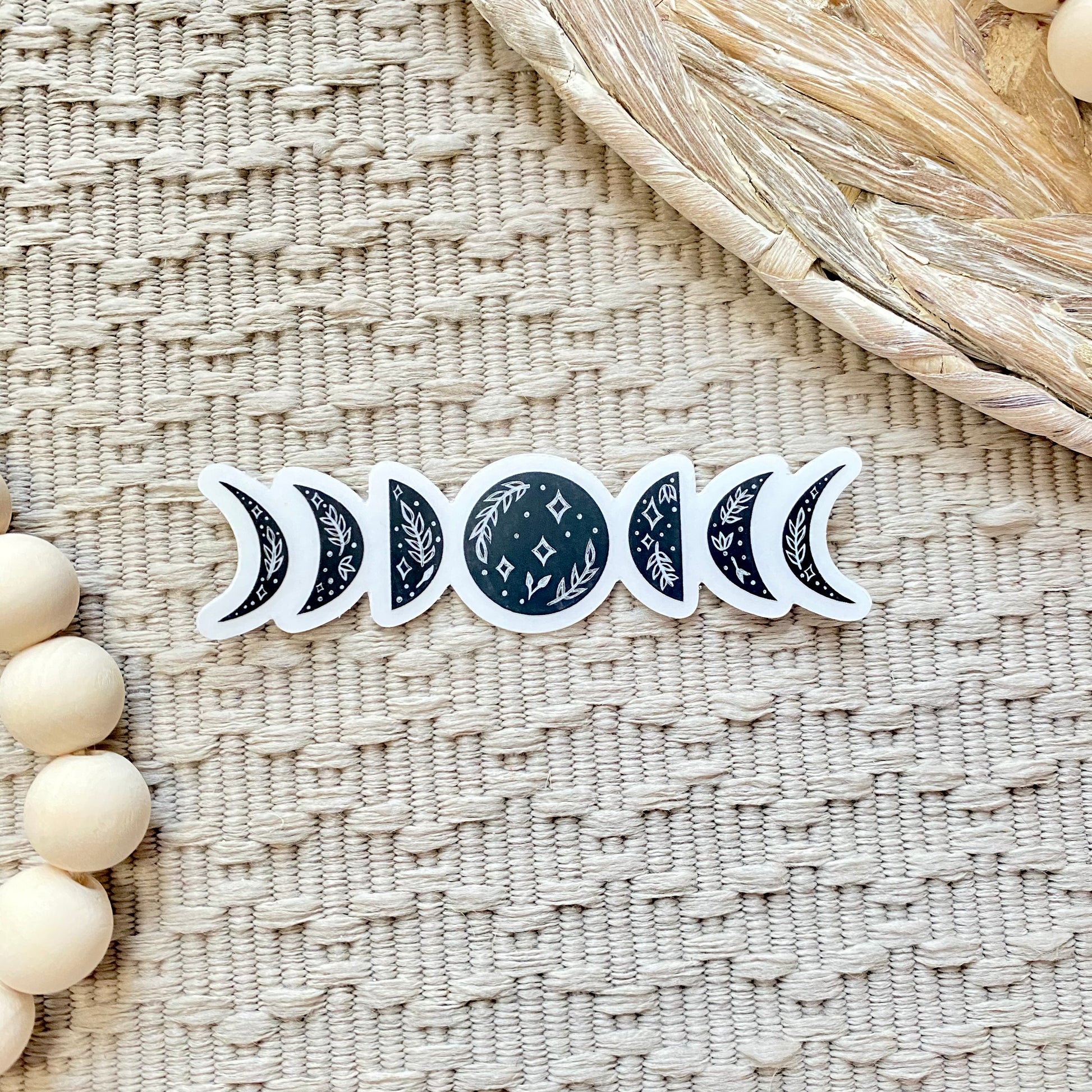 Set of black floral moon phase stickers on a textured woven surface with wooden beads.