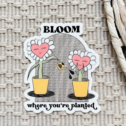 Sticker of two cartoon flowers watering one another and text saying "bloom where you're planted"