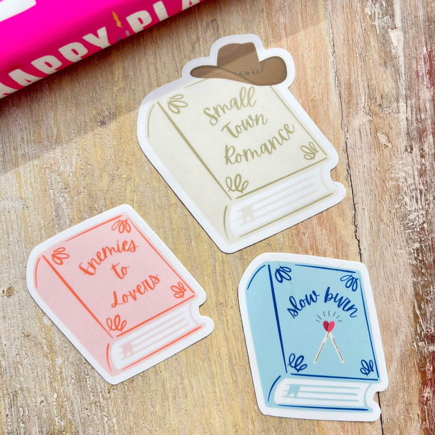 Three book-themed stickers on a wooden surface with a pink book in the background.