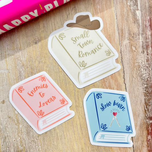 Three book-themed stickers on a wooden surface with a pink book in the background.