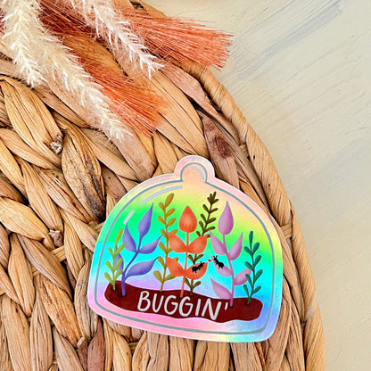 Holographic sticker of terrarium with plant designs and 'BUGGIN' text on a woven surface