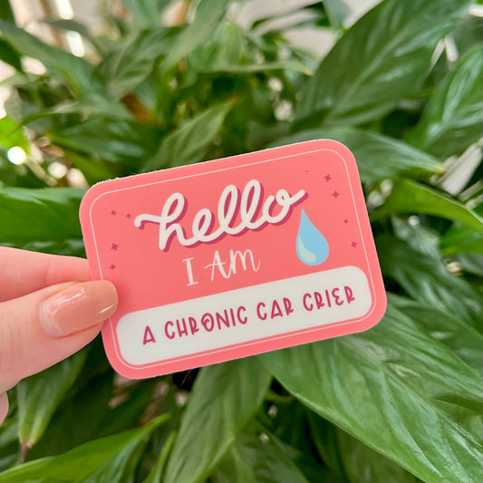 Pink sticker with text 'Hello I am a chronic car crier' held in front of green leaves