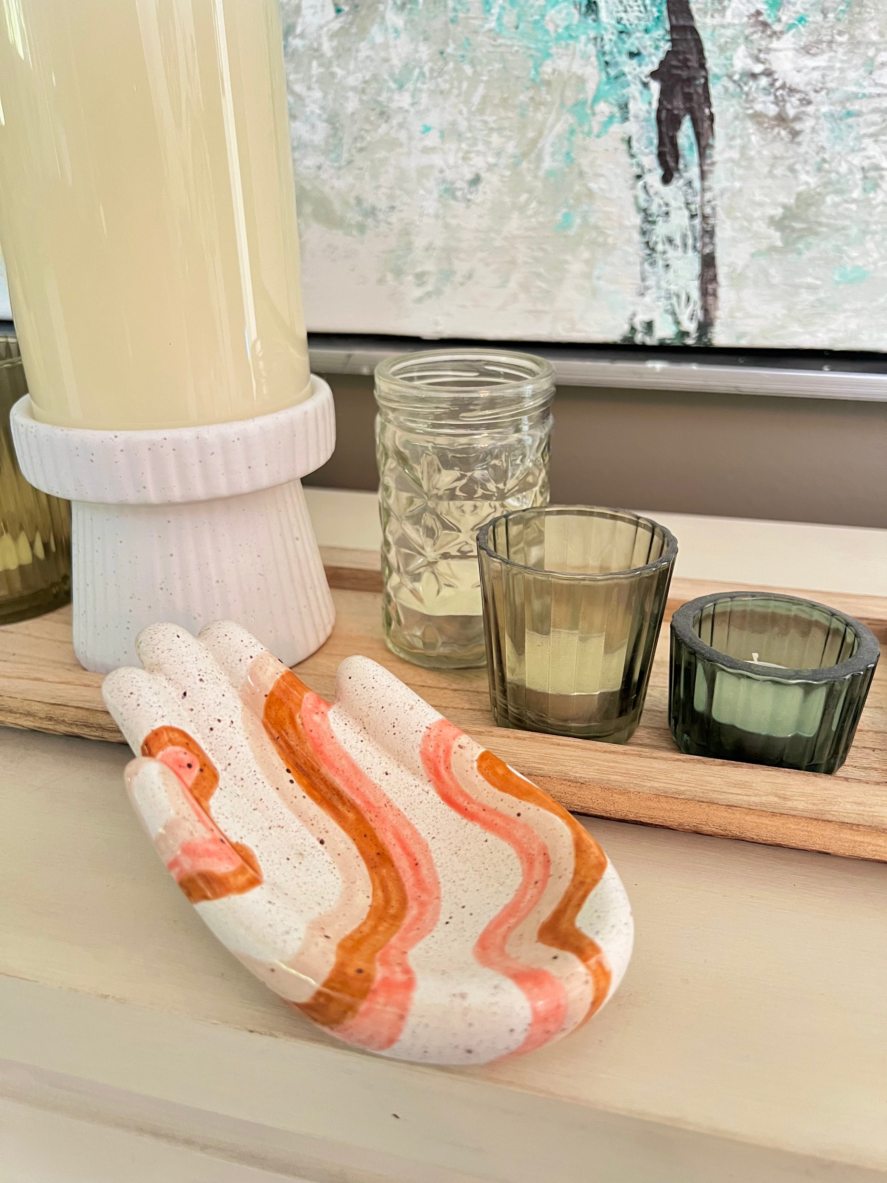 Decorative ceramic hand with wavy pattern on a surface with candles and jars.