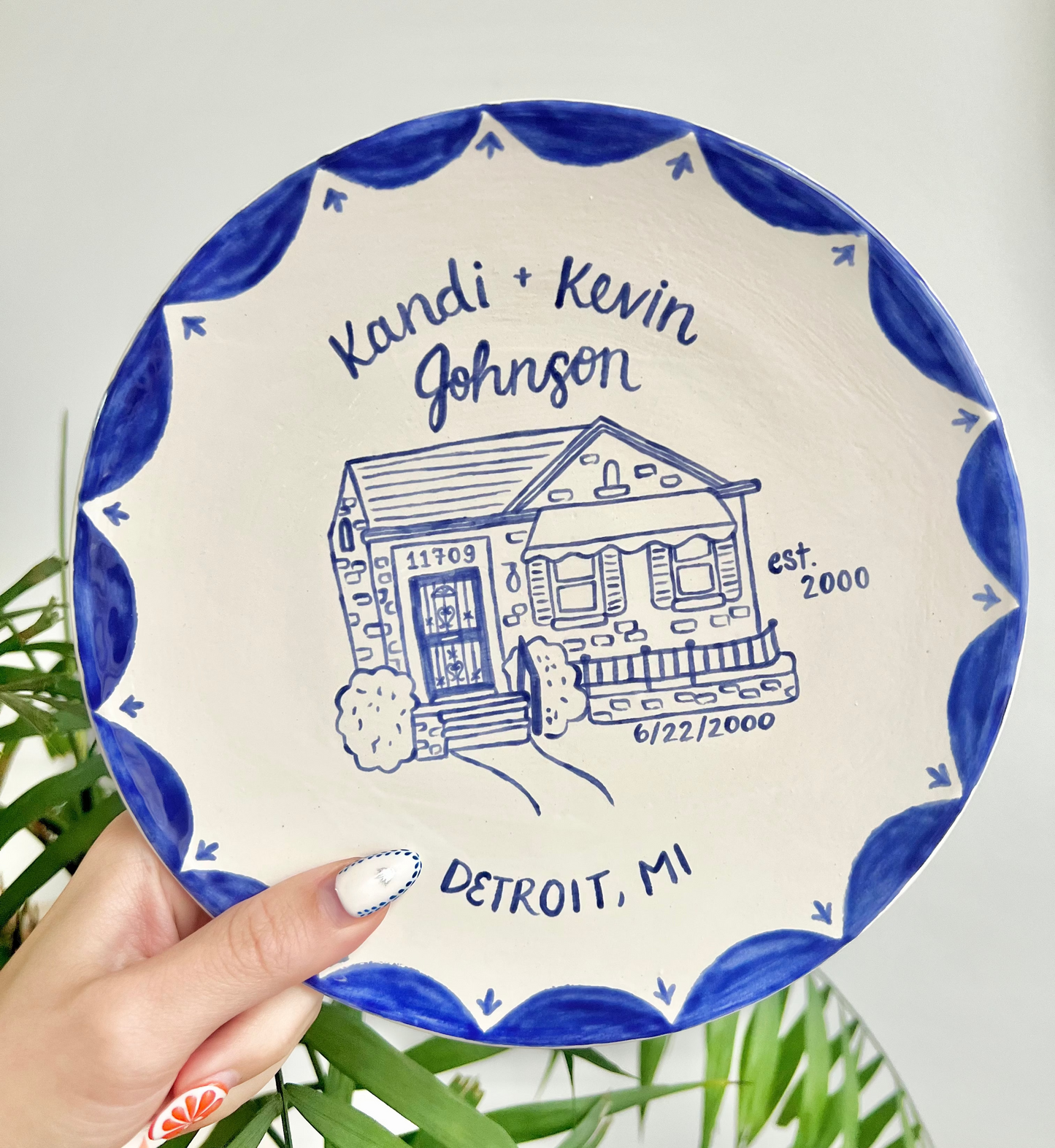 Decorative ceramic plate with house illustration and text held by a hand against a white background