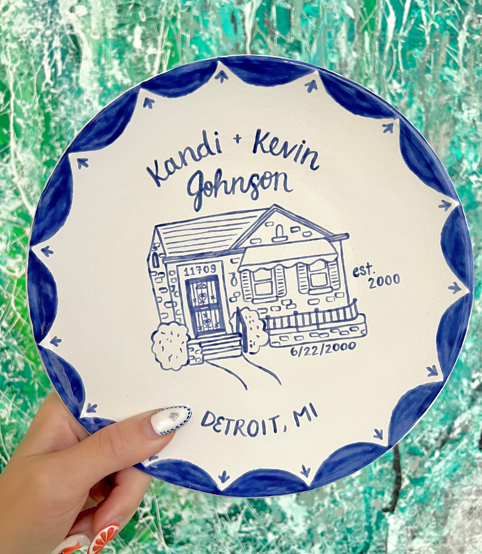 Decorative ceramic plate with house illustration and text, held against a textured green background