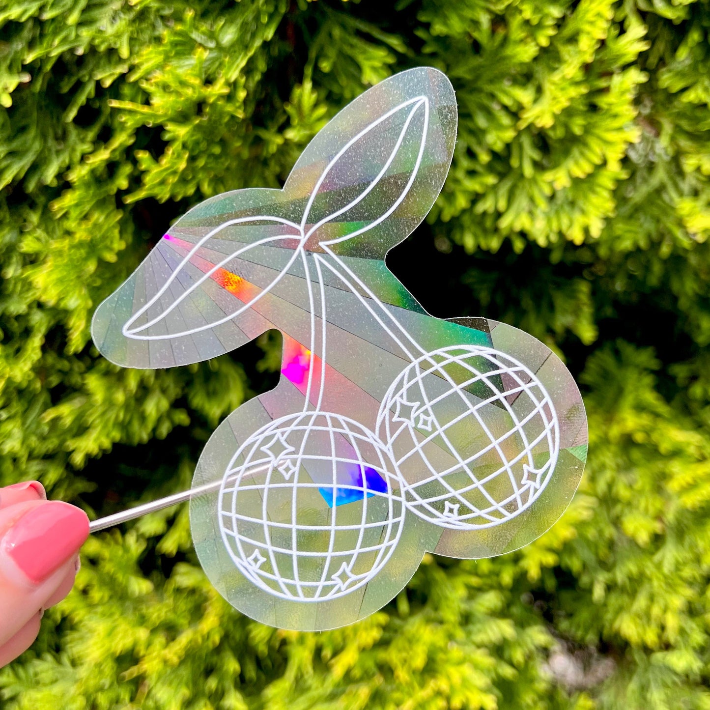 Holographic cherry-shaped suncatcher sticker held against a green leafy background