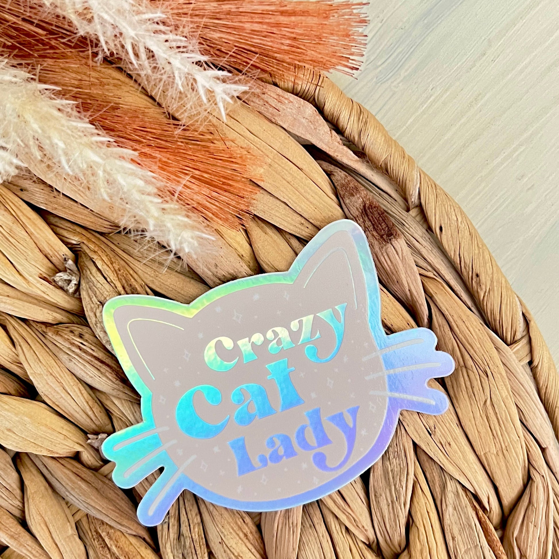 Holographic 'Crazy Cat Lady' sticker on a woven surface with dried plants.