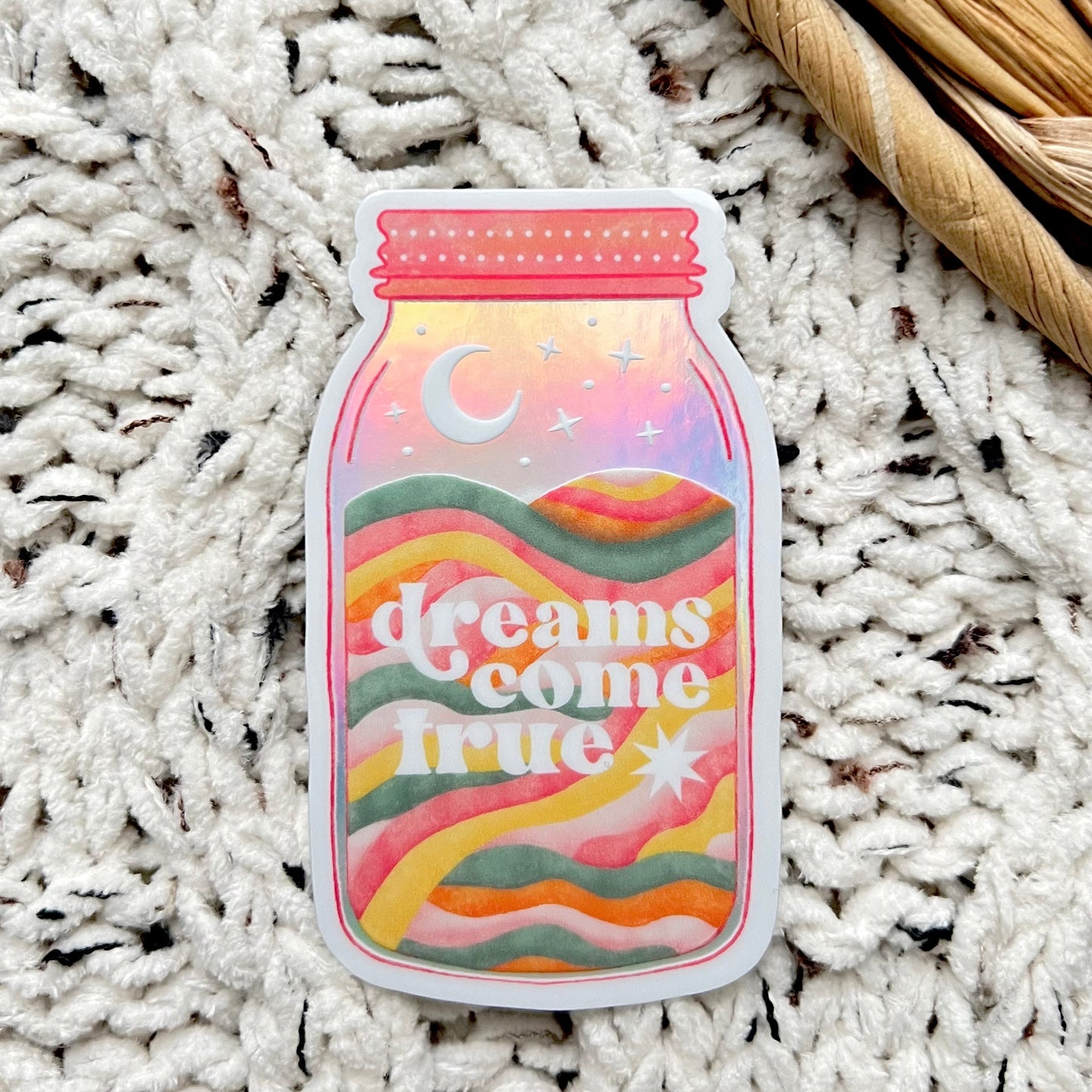 Holographic sticker with 'dreams come true' text written on a mason jar with a moon inside sitting on a textured white surface