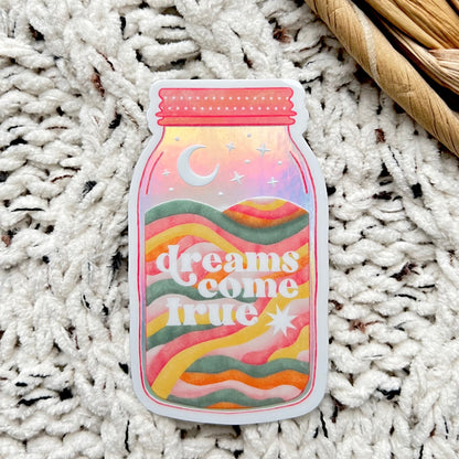 Holographic sticker with 'dreams come true' text written on a mason jar with a moon inside sitting on a textured white surface