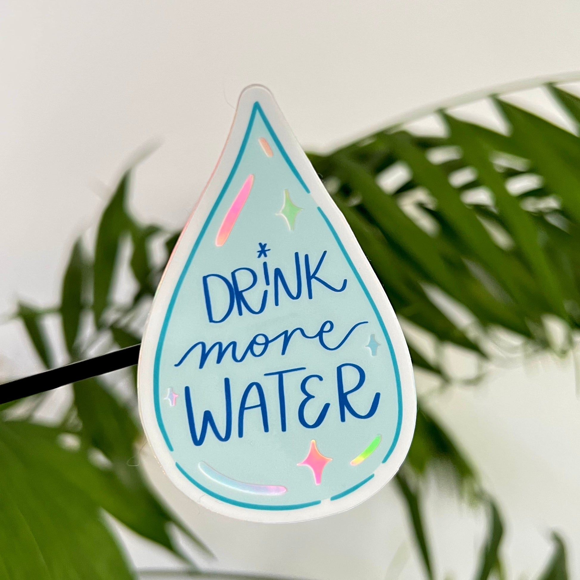 Sticker shaped like a water droplet with 'Drink more water' text, placed against a leafy green background.