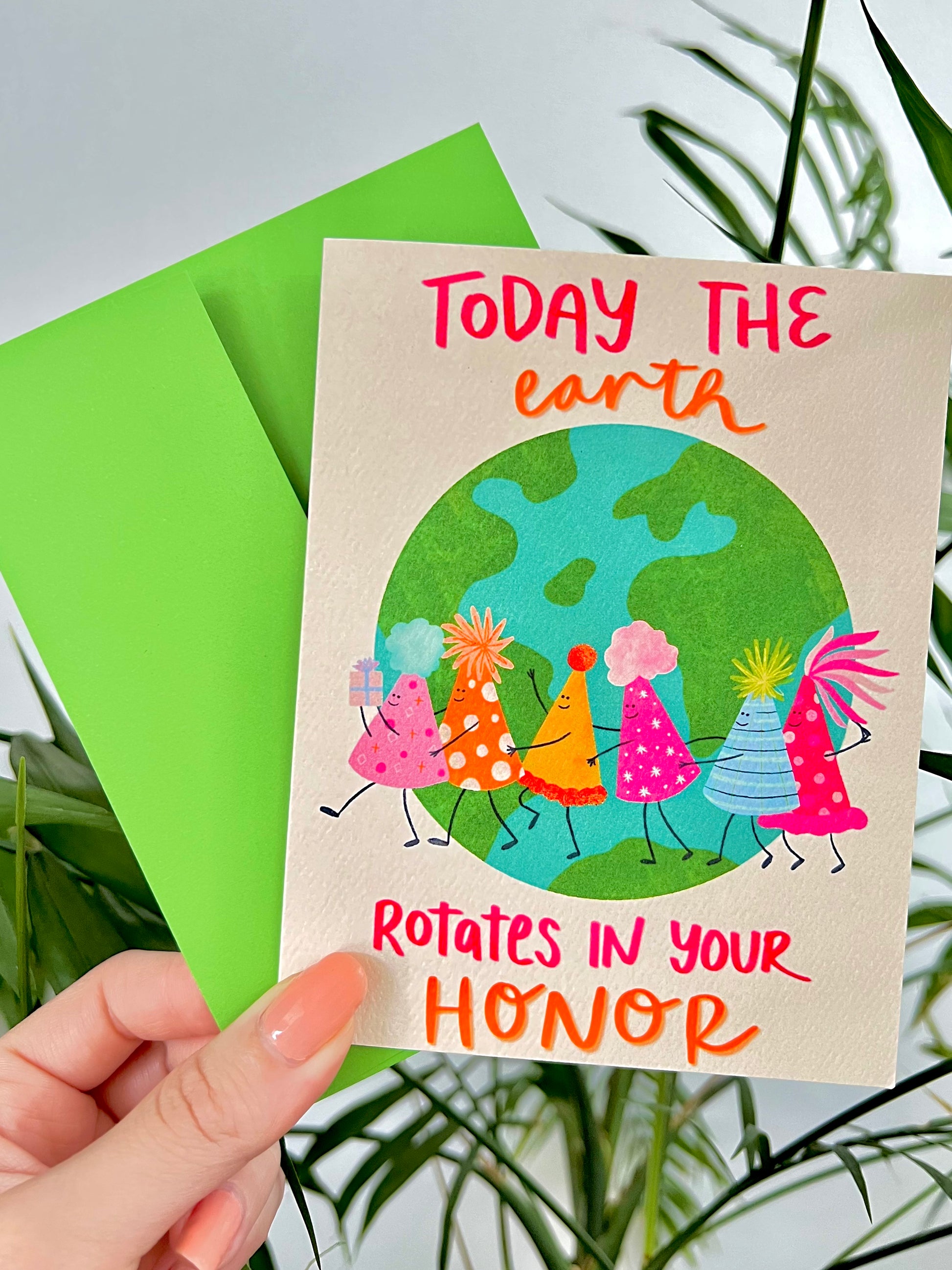 Card with cartoon characters of birthday hats around the Earth and text 'Today the earth rotates in your honor' held by a hand with a green envelope.
