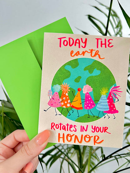 Card with cartoon characters of birthday hats around the Earth and text 'Today the earth rotates in your honor' held by a hand with a green envelope.