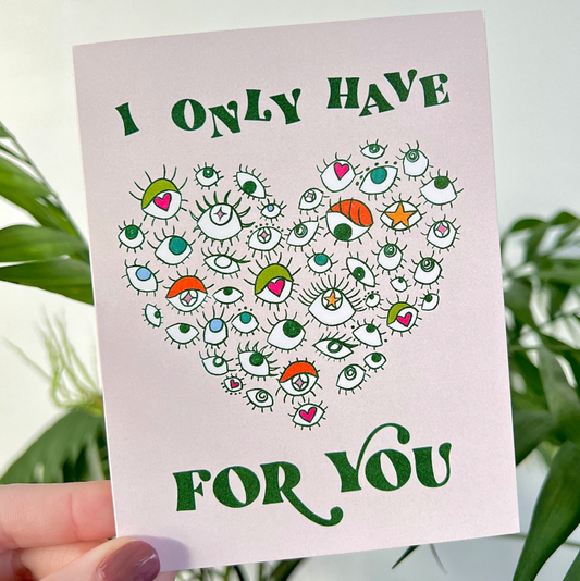 Card with a heart design made of eyes and text that says "I only have eyes for you" held by a hand in front of green plants