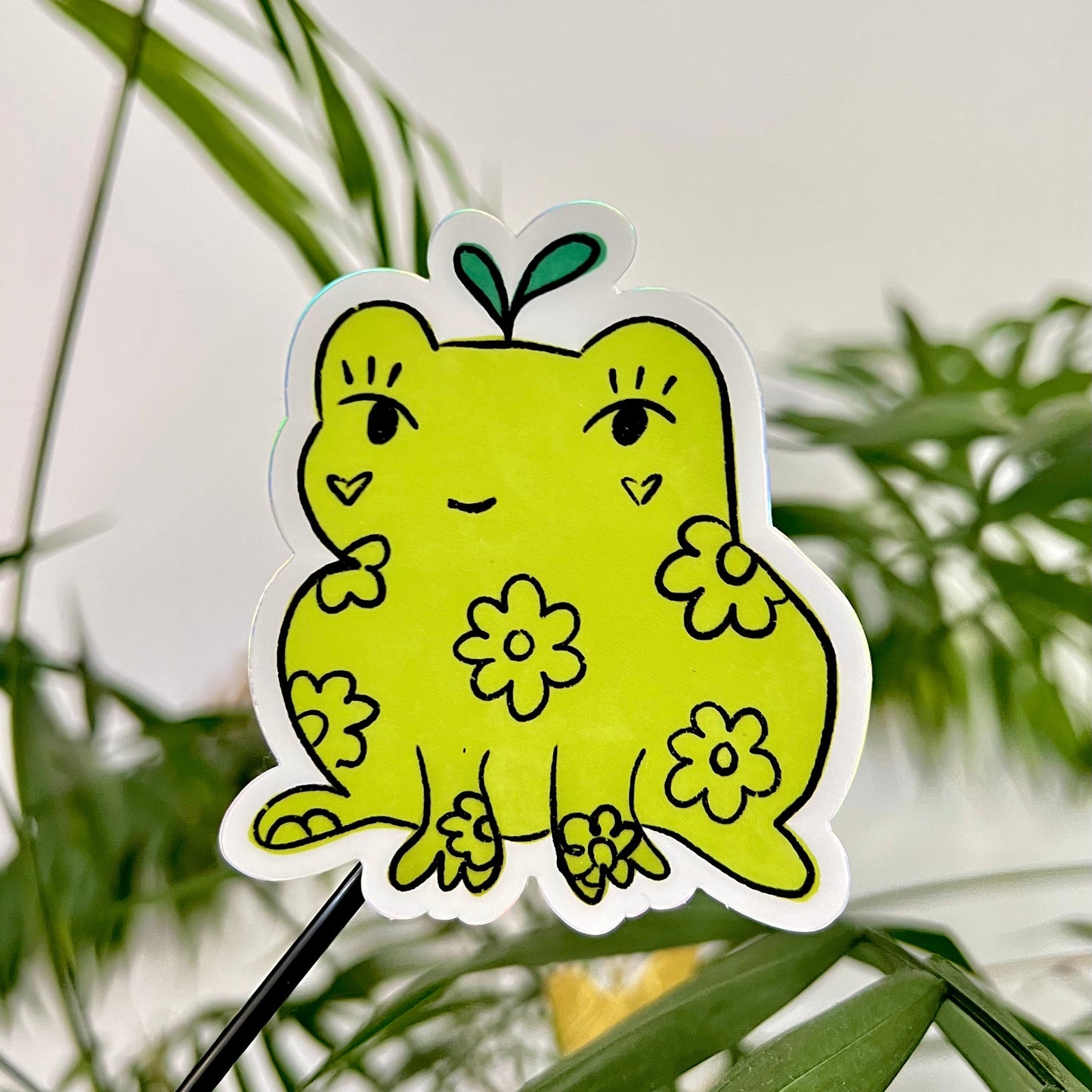 Green frog-shaped sticker with floral designs against a green leafy background