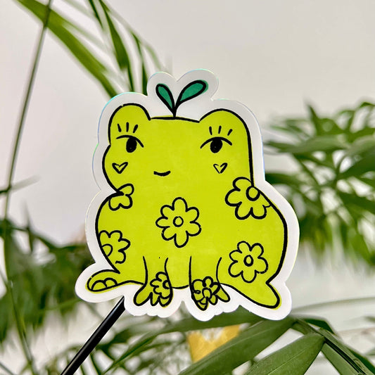 Green frog-shaped sticker with floral designs against a green leafy background