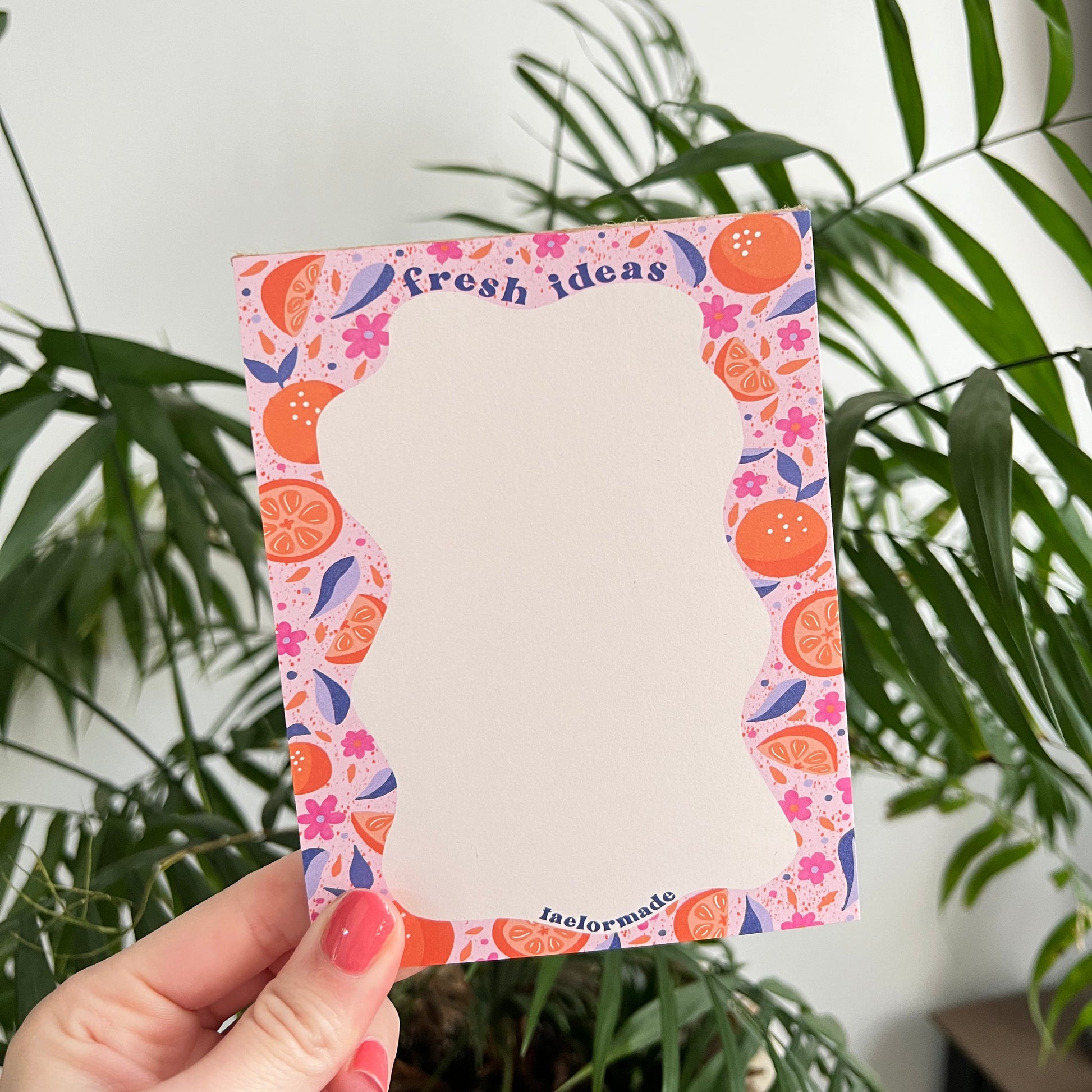 Notepad with colorful fruit border held by a hand in front of green leaves