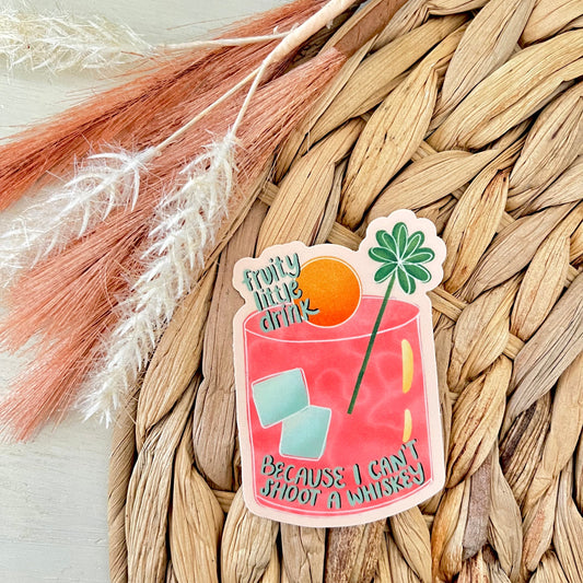 Sticker of a pink drink with text saying "fruity little drink because I can't shoot a whiskey" on a woven mat with dried plants.