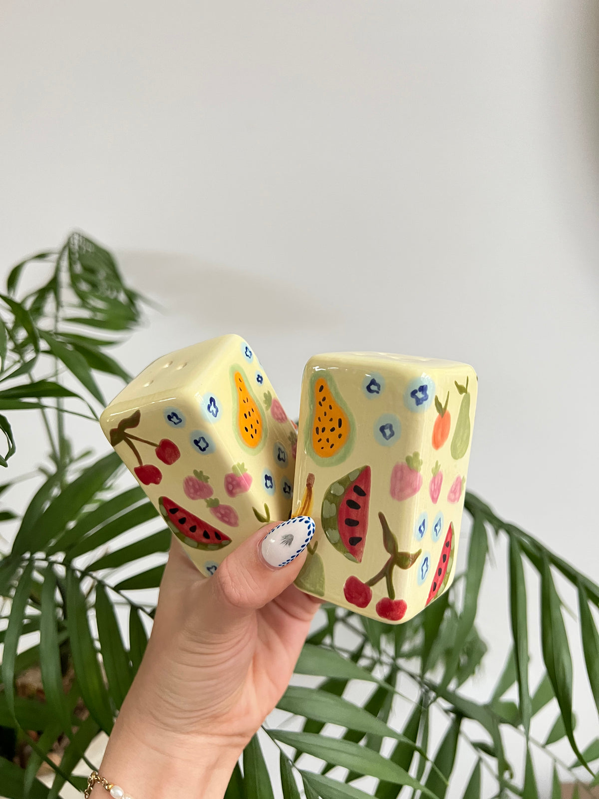Hand holding two ceramic salt and pepper shakers with fruit patterns against a light background