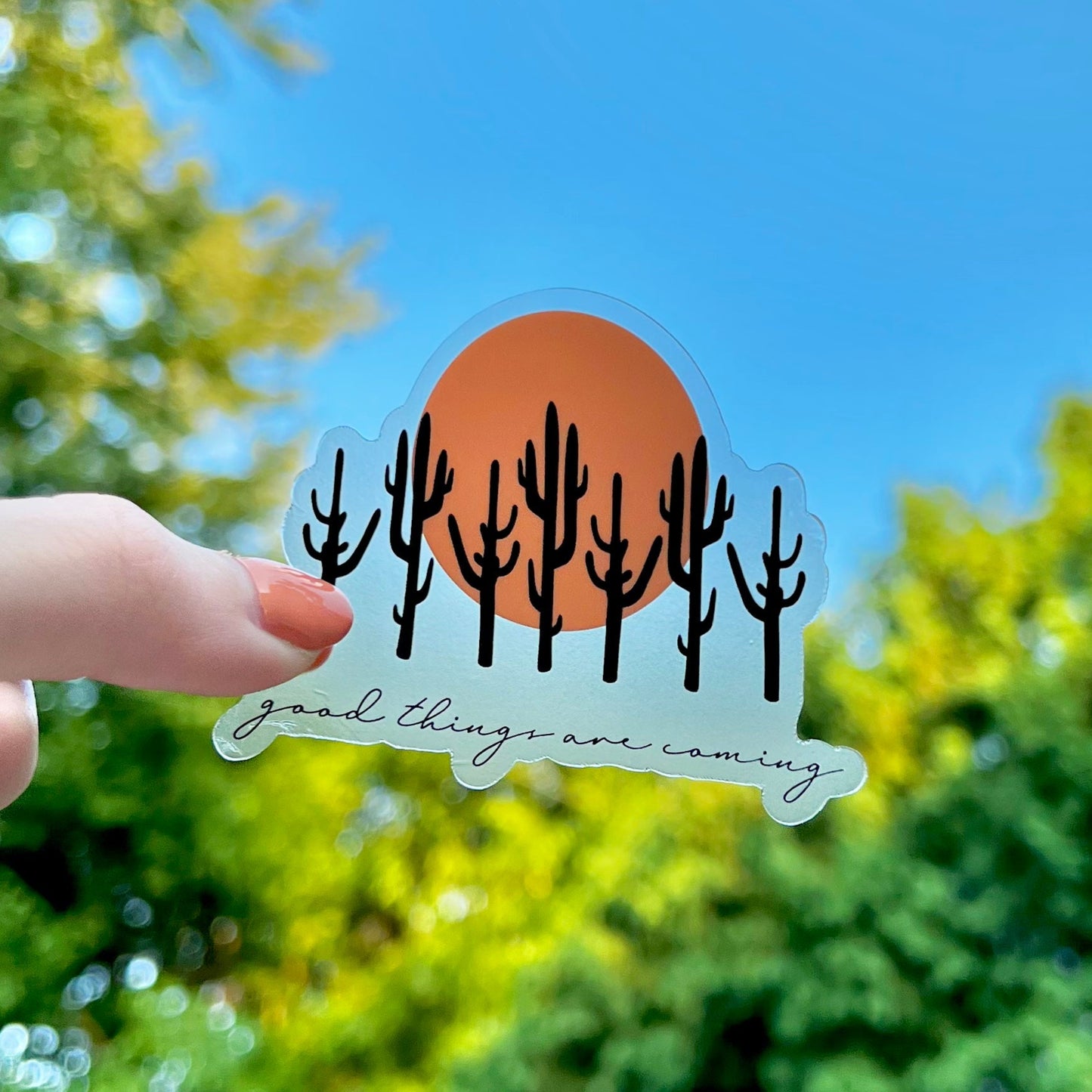 Hand holding a sticker with cacti and 'good things are coming' text against a blue sky and green foliage background.