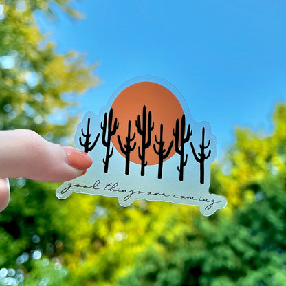 Hand holding a sticker with cacti and 'good things are coming' text against a blue sky and green foliage background.