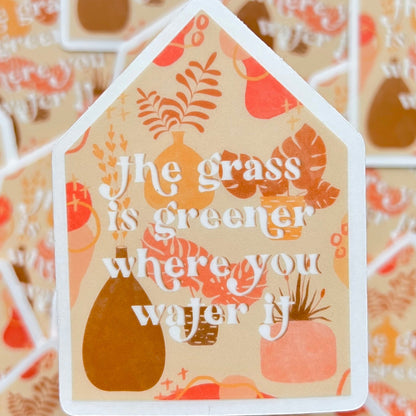 Decorative sticker shaped like a house with 'the grass is greener where you water it' text and illustrations on a patterned background.