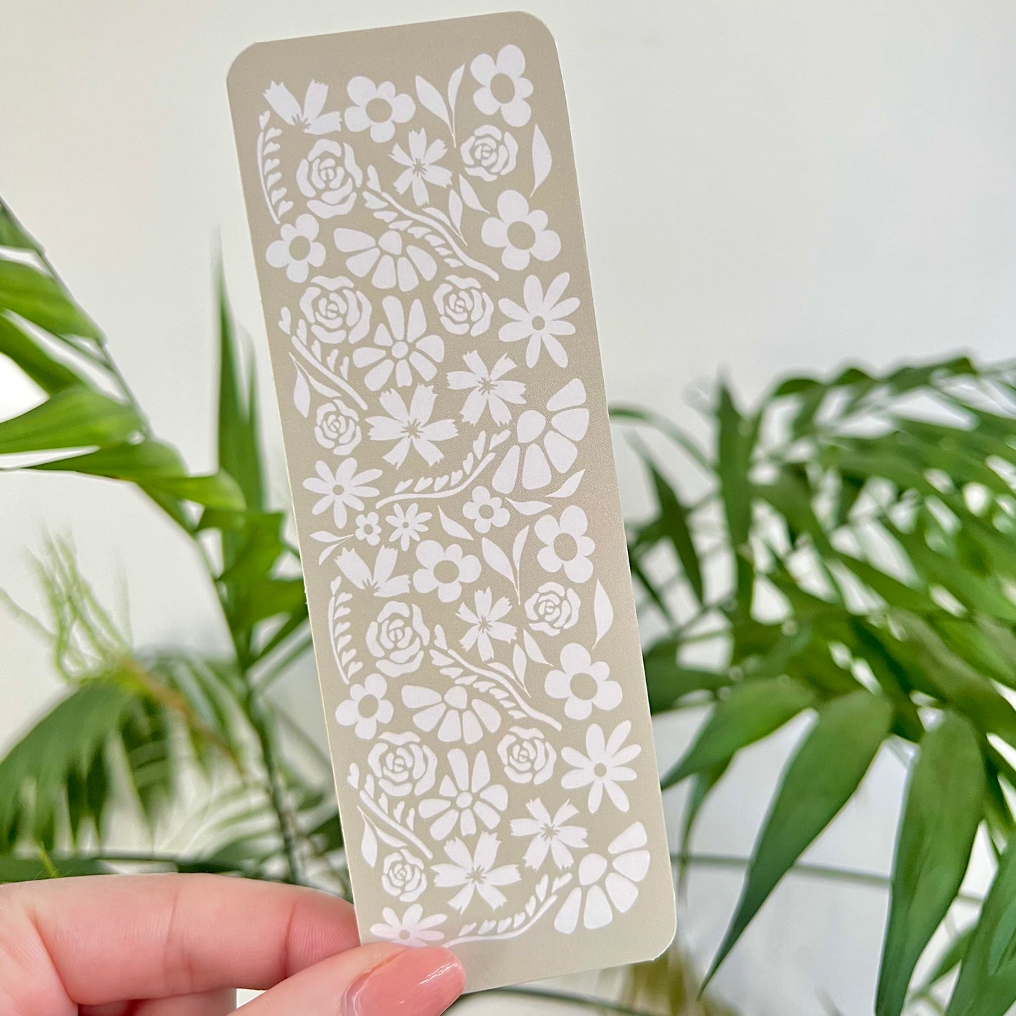 Hand holding a floral patterned bookmark against a blurred green plant background