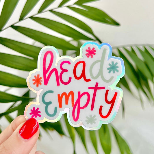 Holographic sticker with 'head empty' text held by a hand against a leafy background