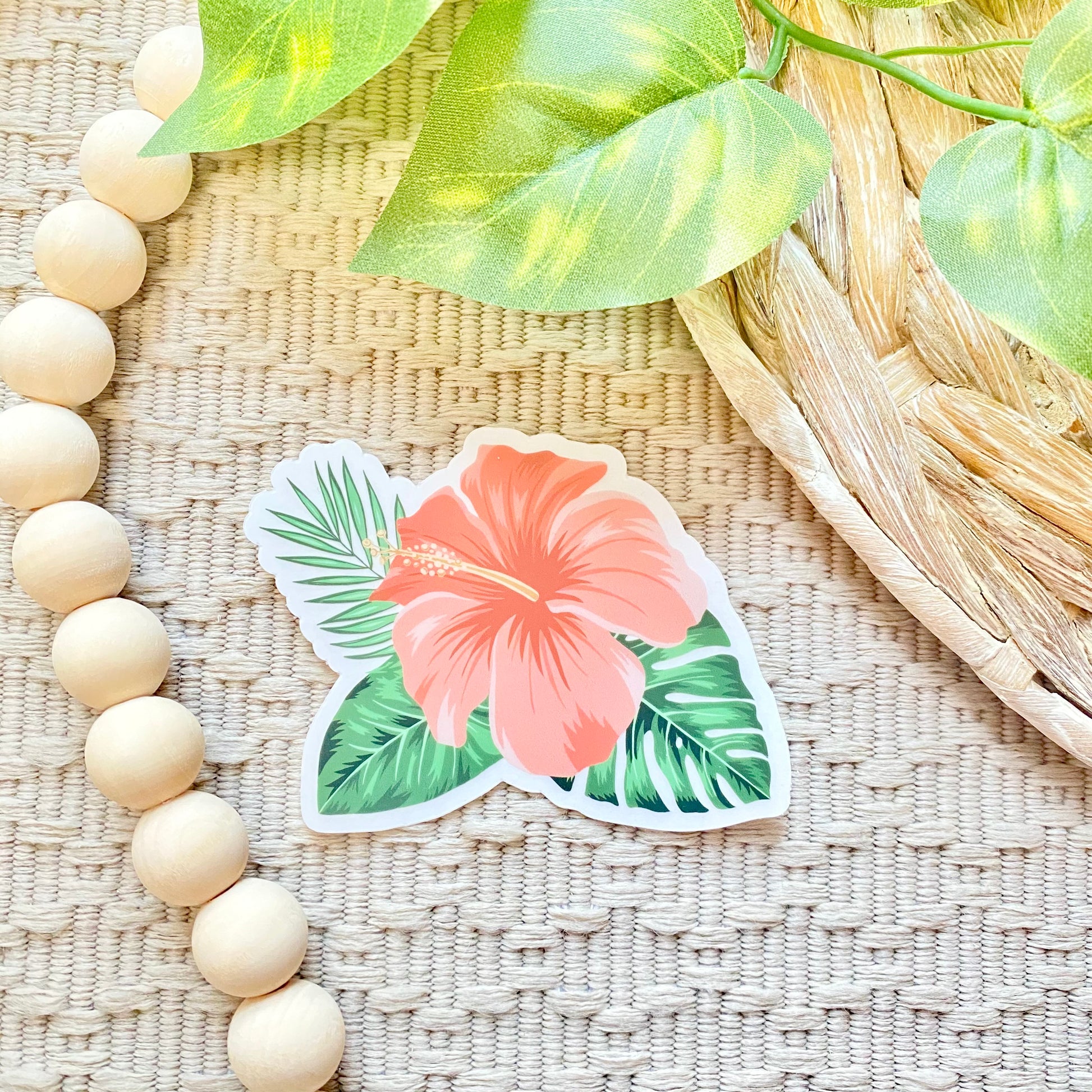 Floral sticker with pink flower and green leaves on a woven surface with beads and leaves.
