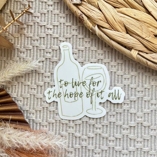 Sticker with wine bottle and glass design and text saying "to live for the hope of it all" in cursive on a textured surface