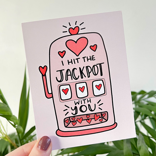 Card with slot machine illustration and 'I hit the jackpot with you' text, held against a natural background.