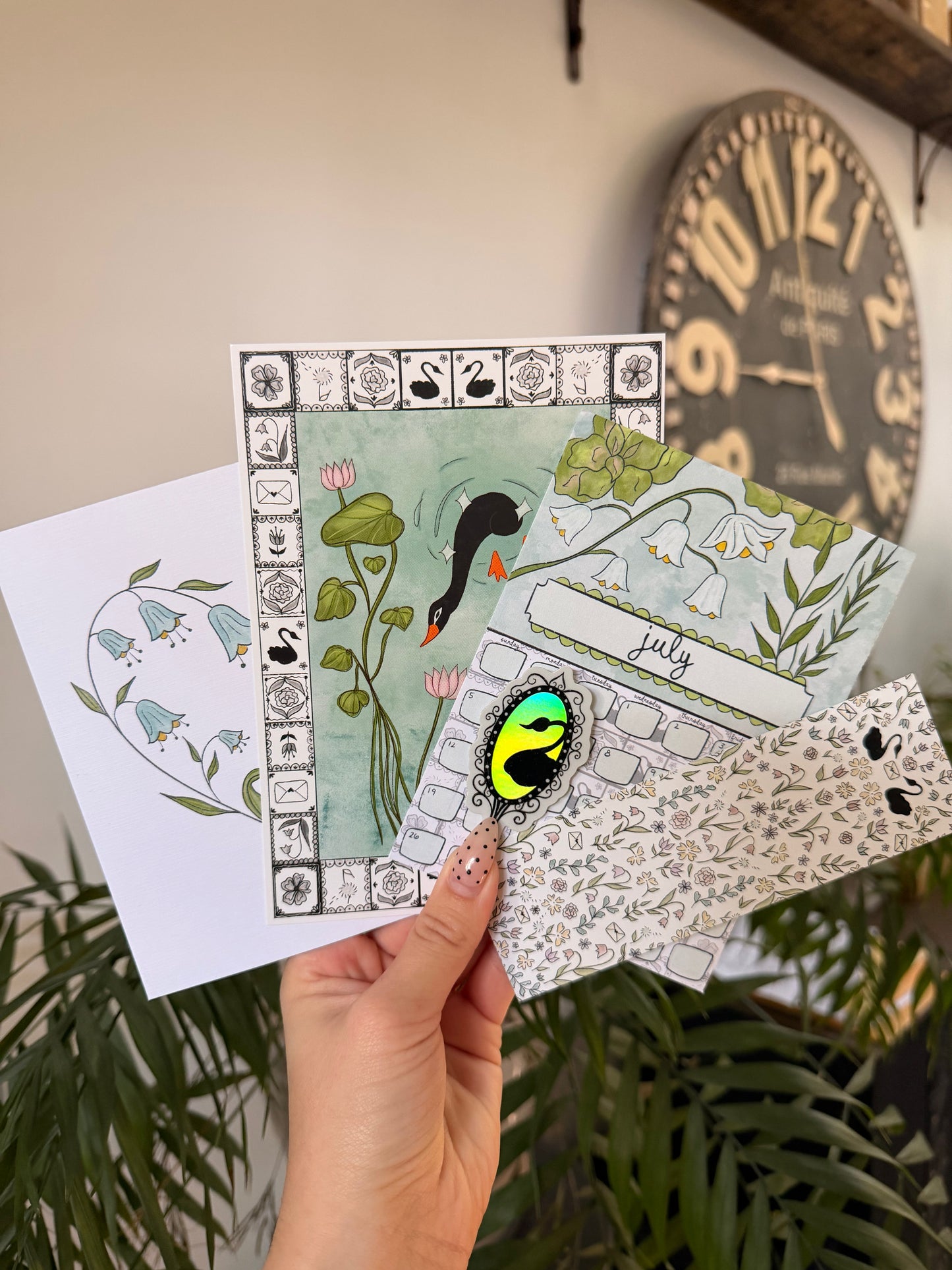 Hand holding three decorative cards with nature-themed designs, a clock in the background.