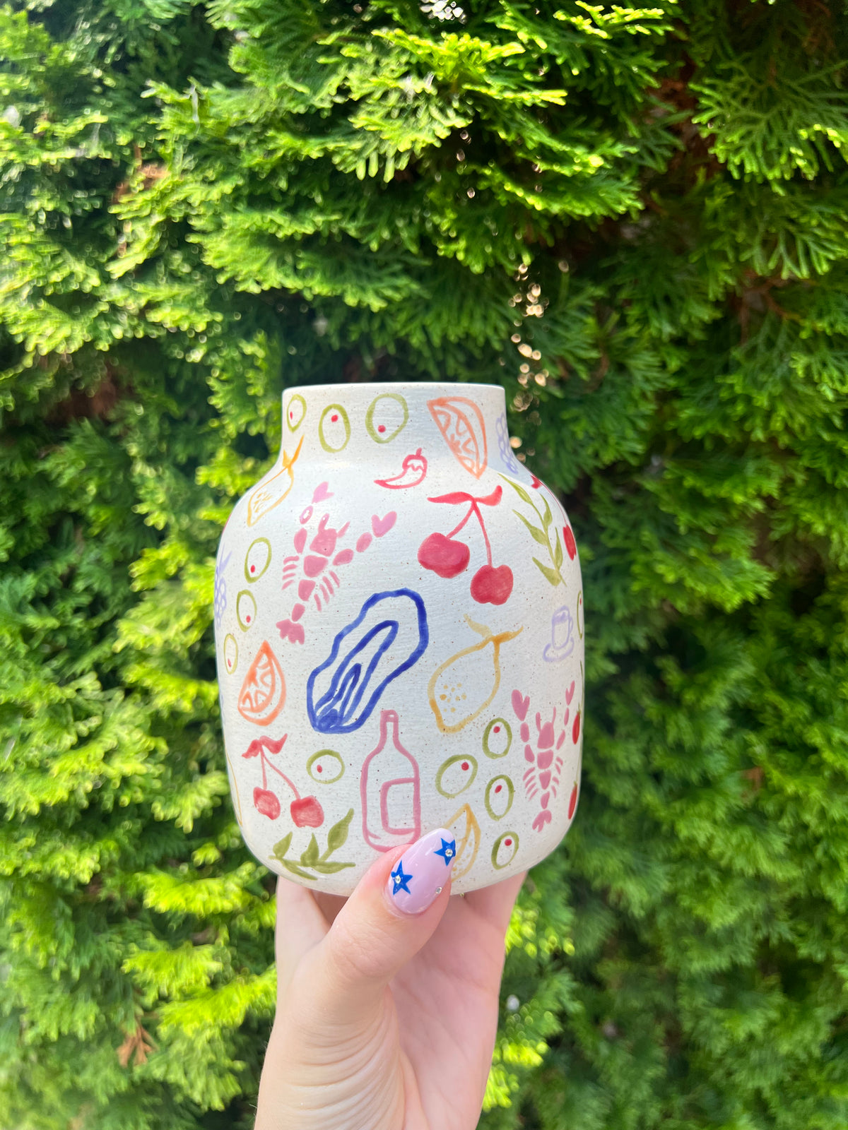 Hand holding a ceramic vase with colorful drawings against a green bush background