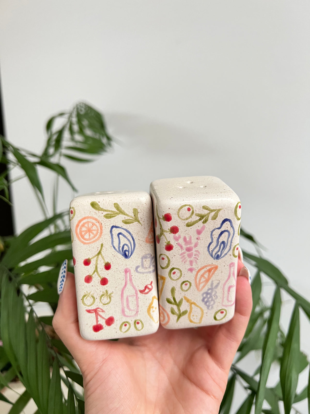 Two ceramic salt and pepper shakers with colorful designs held by a hand against a plant background.
