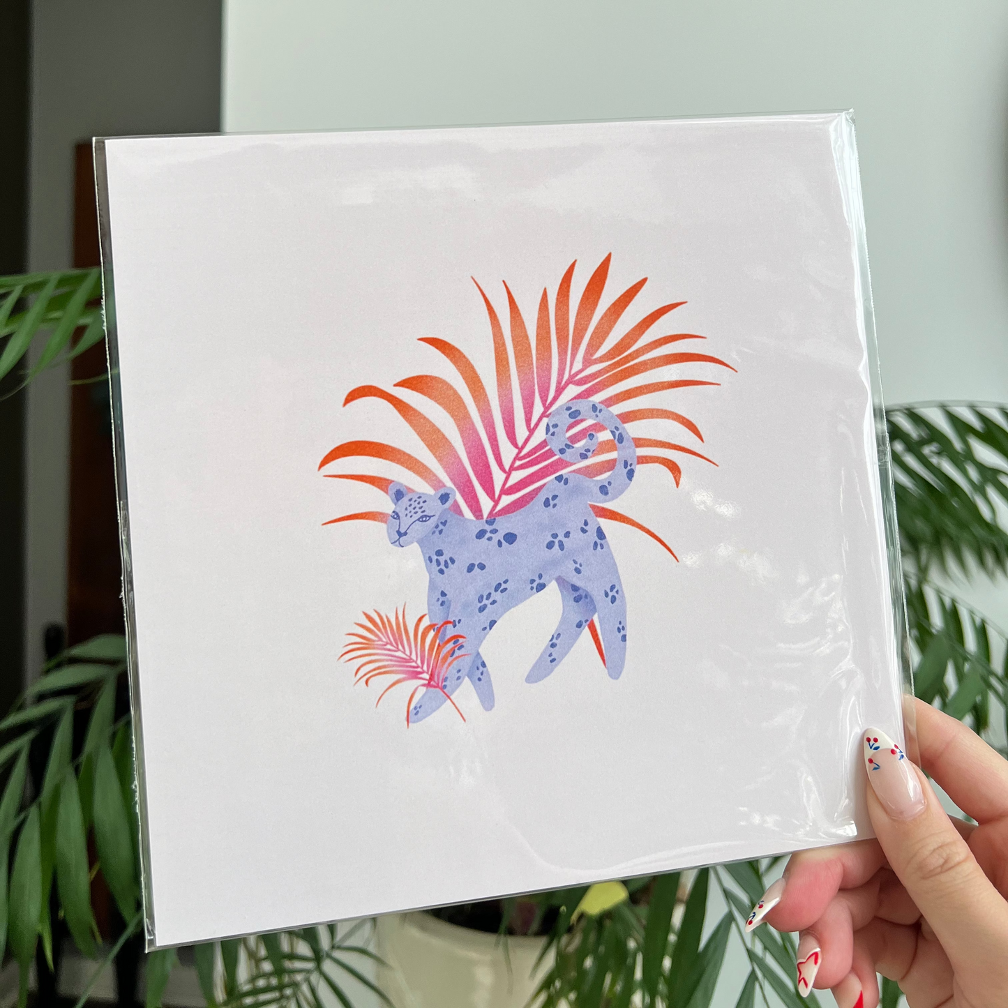 Hand holding a white square with a colorful design of a leopard and plants against a light background.