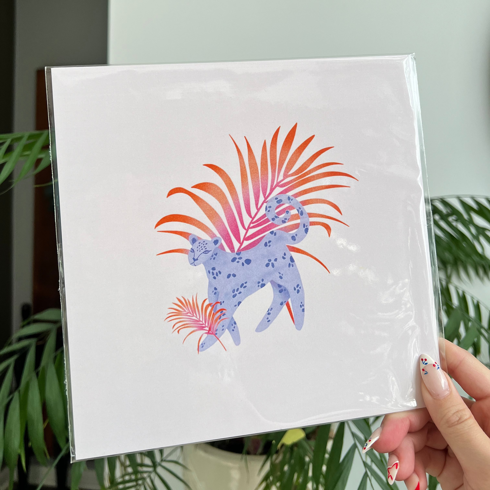 Hand holding a white square with a colorful design of a leopard and plants against a light background.